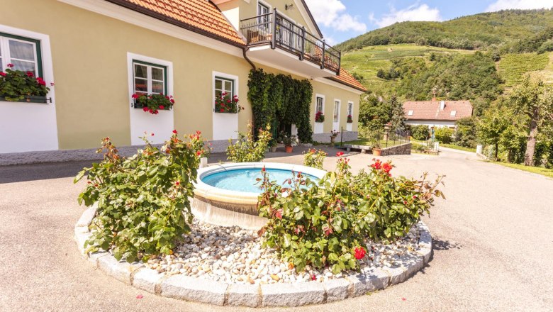 Entrance to a winery with fountain and flowers, surrounded by hills.
