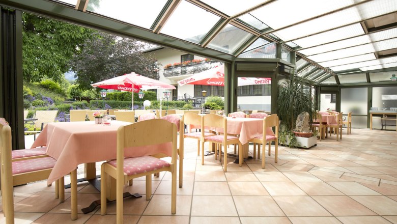Bright restaurant with glass roof, pink tablecloths and view of the garden with parasols.