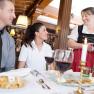 A couple sits at a set table in a restaurant while a waitress in traditional costume serves them with a smile.