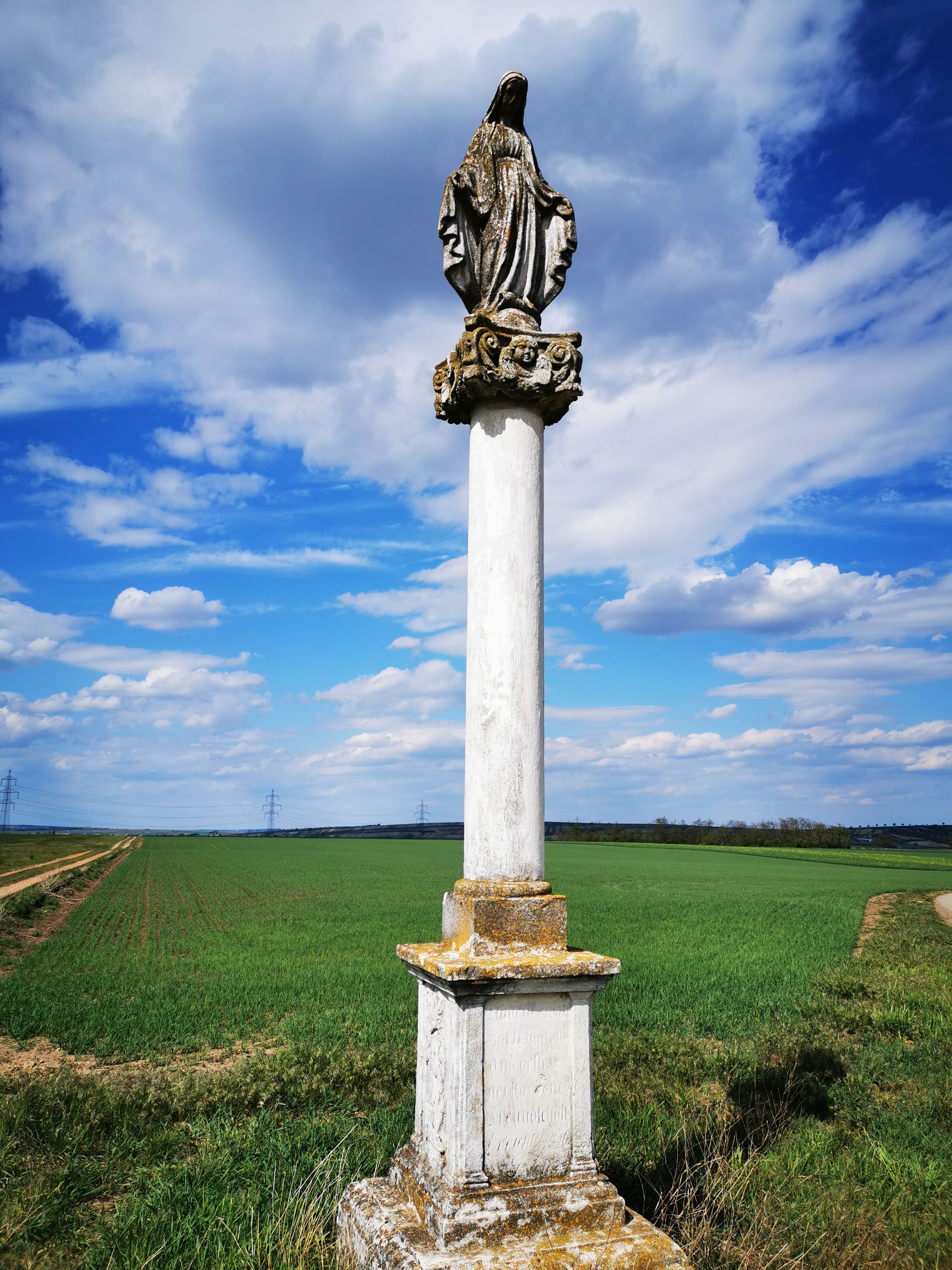 A statue on a pillar in a rural landscape under a blue sky with clouds.