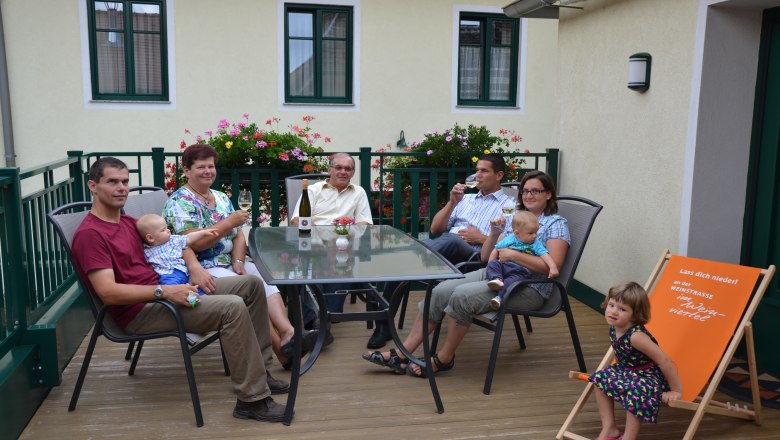 Krottendorfer family, © Familie Krottendorfer A family is sitting on a terrace at a table with drinks. Flowers and a building can be seen in the background.