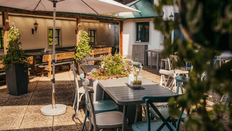 A sunny terrace with tables, chairs and plants under a parasol.