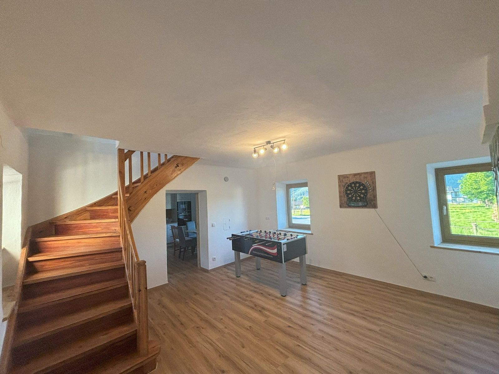 A common room with table football table, dartboard on the wall, wooden staircase and two windows.