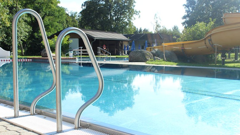 An outdoor pool with slide and ladder, surrounded by trees and a building in the background.