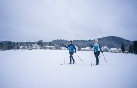 Two people cross-country skiing on a snow-covered surface in front of a village and wooded hills.