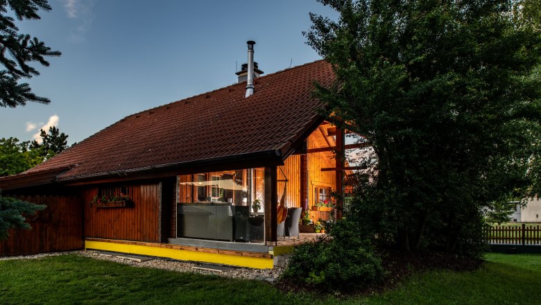 Exterior view of a wooden house with large windows and a garden surrounded by trees.