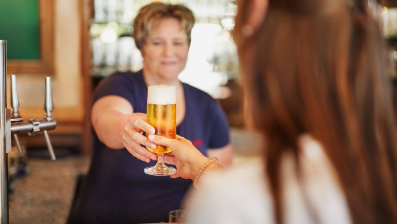 A woman hands a guest a freshly tapped beer in a glass across the bar.