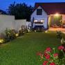 An illuminated courtyard at night with a manicured lawn, flower beds and a small building in the background.