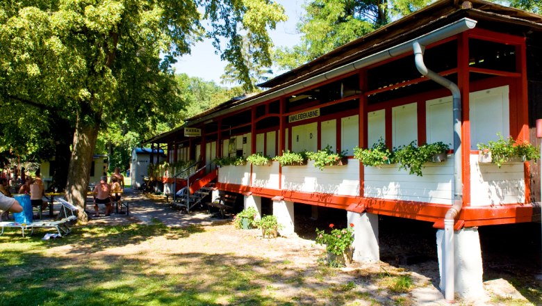 A red building with changing rooms and plants in front of it, surrounded by trees and people on benches.