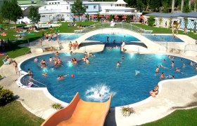 Outdoor swimming pool with bridge and slide, lots of people swimming and sunbathing.