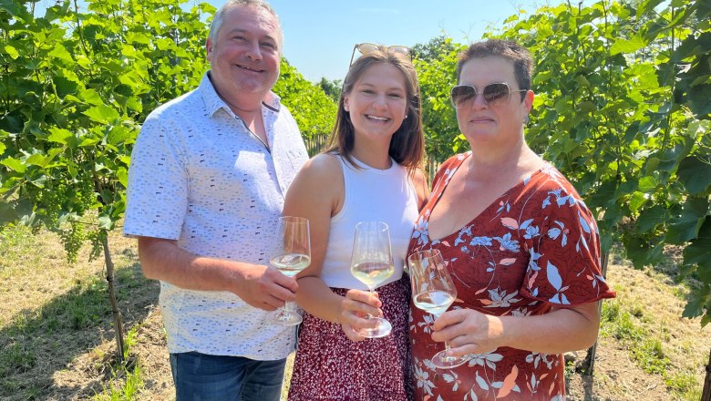 Familie Heimel mit Weingläsern in der Hand. Die Familie steht zwischen Weinreben.