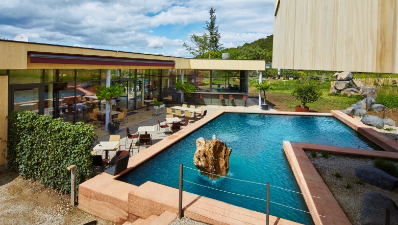 Modern terrace with tables and chairs next to a rectangular pond with rocks in the middle.