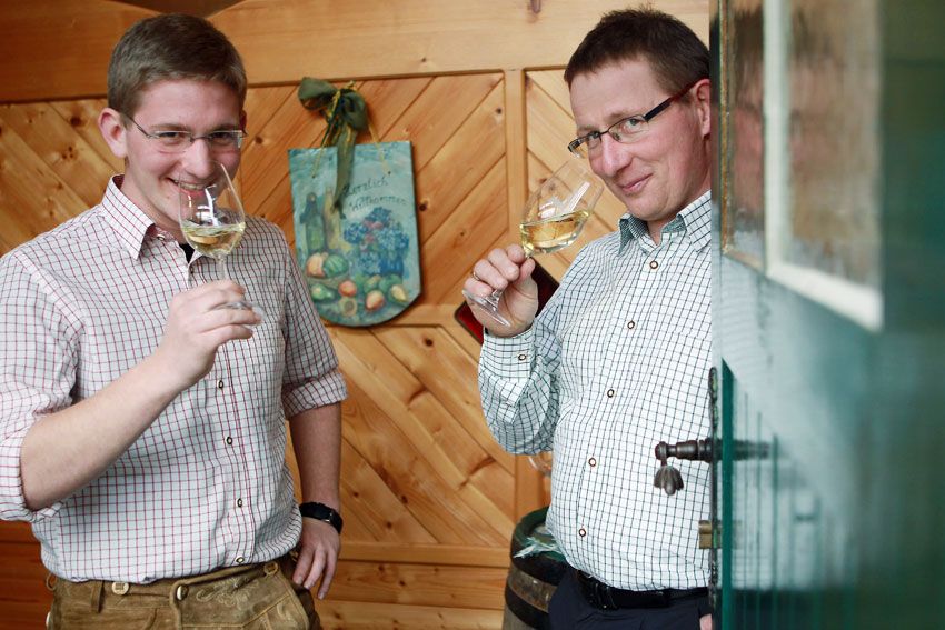 Two men in checked shirts drink white wine in a wood-paneled room.