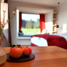 A modern hotel room with a red bedspread, a table with apples and a window with a view of the greenery.