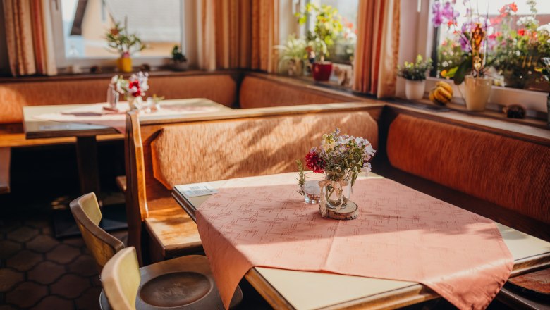Cozy parlor with wooden furniture, tablecloths and flowers on the tables, surrounded by windows with plants.
