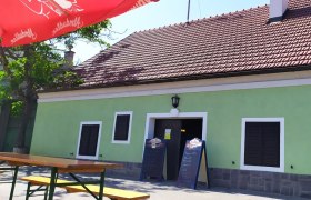 Exterior view of a traditional wine tavern with green paint and red roof tiles. There are wooden benches and a red parasol in front of the building.