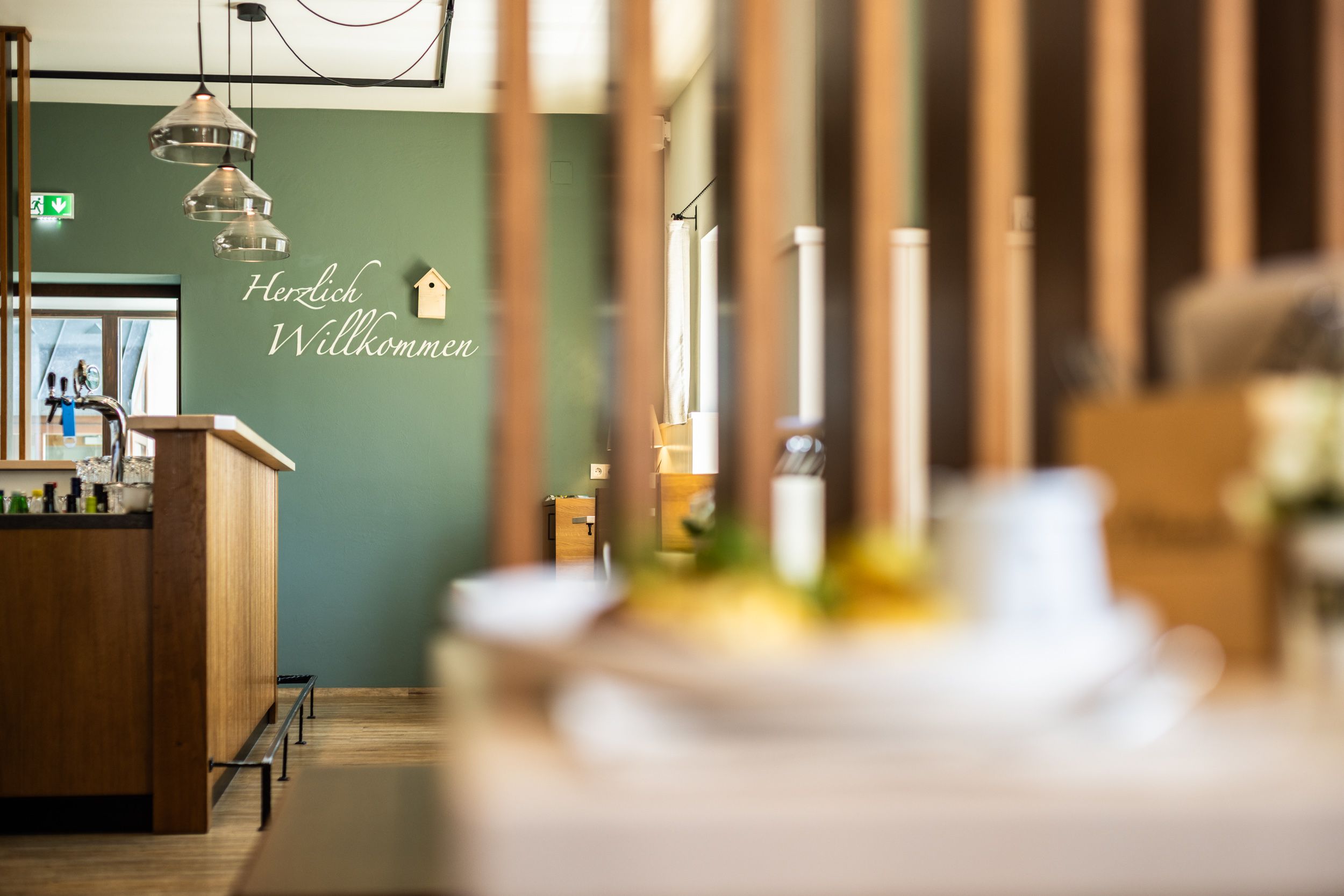 Interior view of a modern restaurant with a green wall and "Welcome" lettering. Lamps hang above the wooden bar and a small birdhouse decorates the wall.
