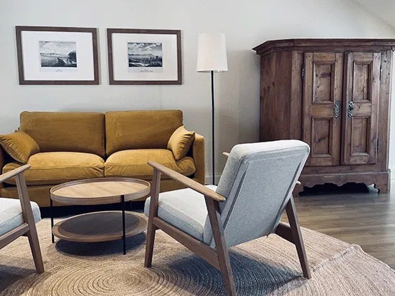 Cozy living room with yellow sofa, two grey armchairs, round wooden table and rustic wooden cupboard.