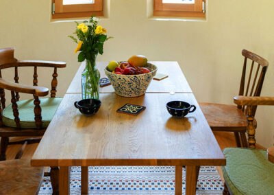 A wooden table with a bouquet of flowers and a bowl of fruit, surrounded by wooden chairs.