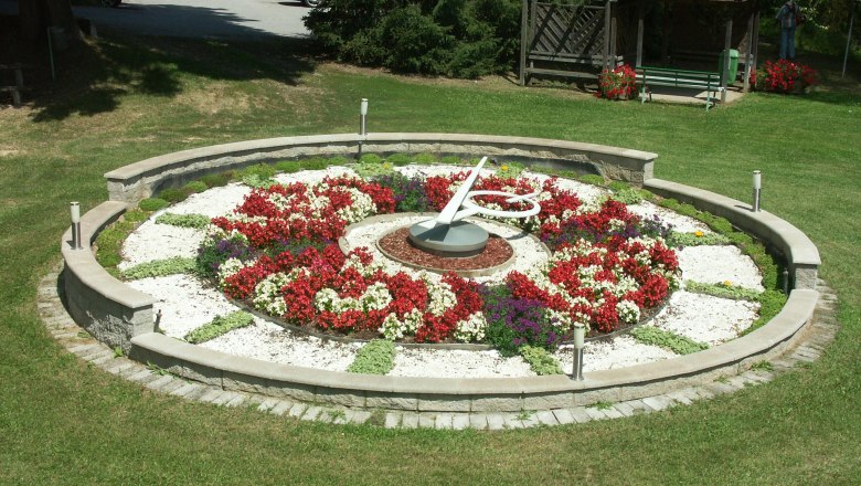 Flower clock in a park with colorful flowers and a pavilion in the background.