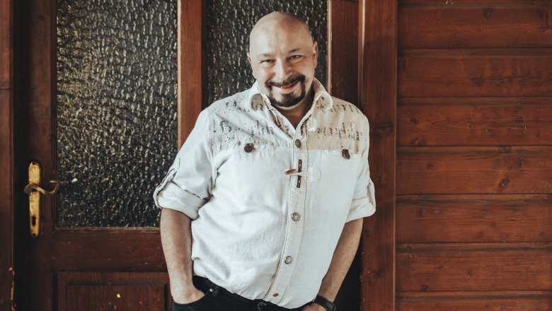 A man in a white shirt leans against a wooden wall, smiling.