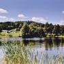 An idyllic campsite with tents on the shore of a lake, surrounded by trees and meadows under a blue sky.