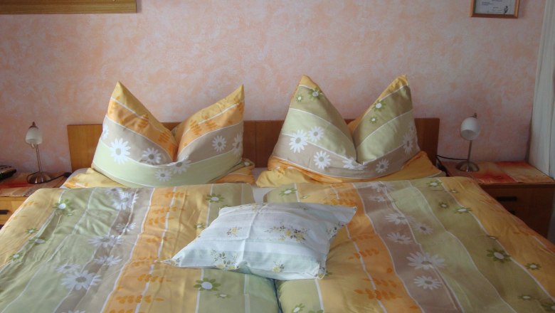 A cozy bedroom with a double bed covered with yellow-green bed linen and floral decor. Two bedside lamps stand next to it.