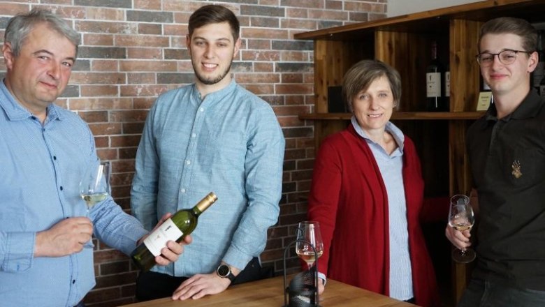Four people stand at a table with wine glasses and a bottle of wine in front of a brick wall.