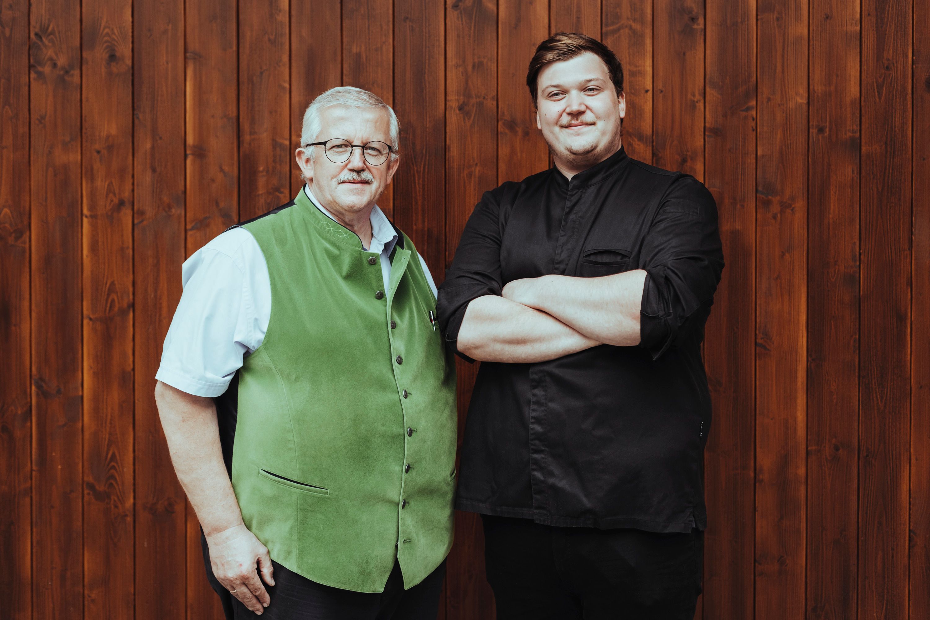 Two men are standing in front of a wooden wall, one wearing a green vest, the other a black shirt.