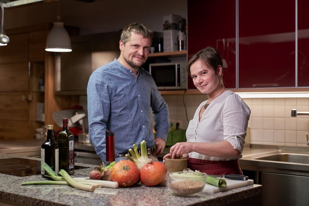 Two people in a kitchen with vegetables and bottles of wine on the worktop.