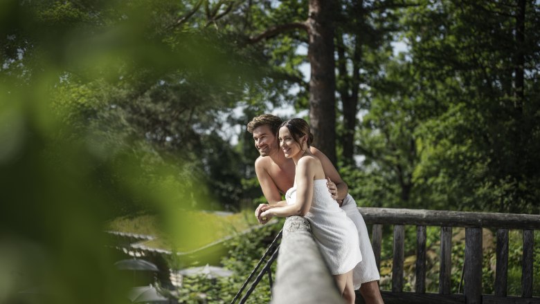 A couple in towels lean against a wooden railing in a green, wooded setting.