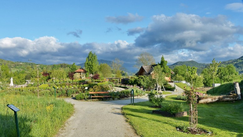 A well-tended garden with paths, trees and flowers, surrounded by green hills and a blue sky with clouds.