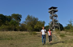 Two hikers in front of a wooden observation tower in a meadow landscape.