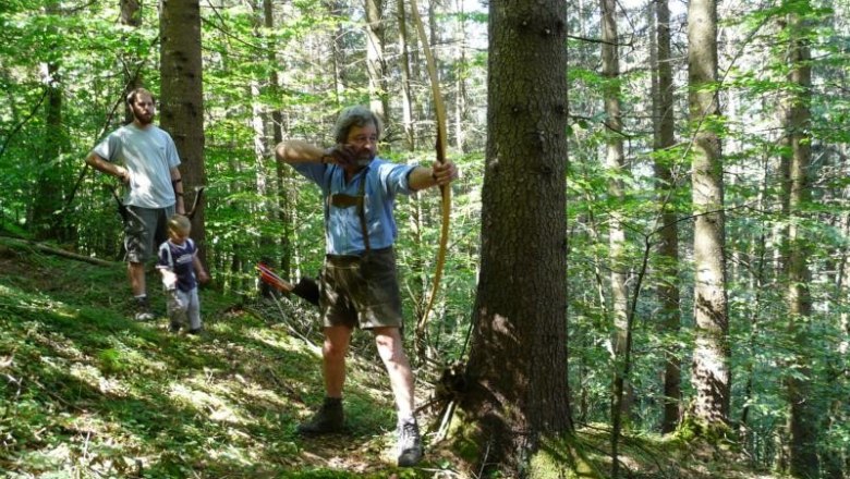 Person shoots with a bow in the forest, two other people in the background.