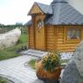 A small, wooden barbecue hut with a gray roof, surrounded by stones and plants.