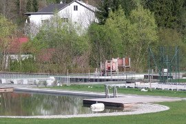Natural swimming pool with jetty, surrounded by trees and a building in the background.