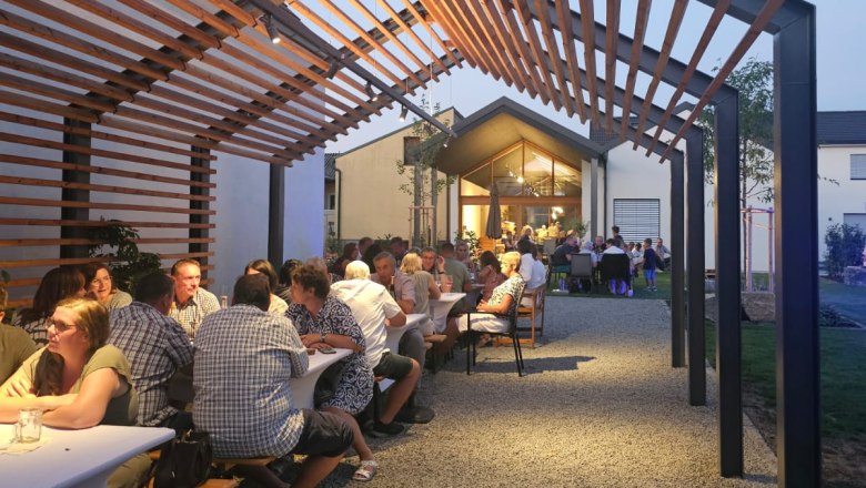 People sit at long tables under a wooden pergola at dusk.