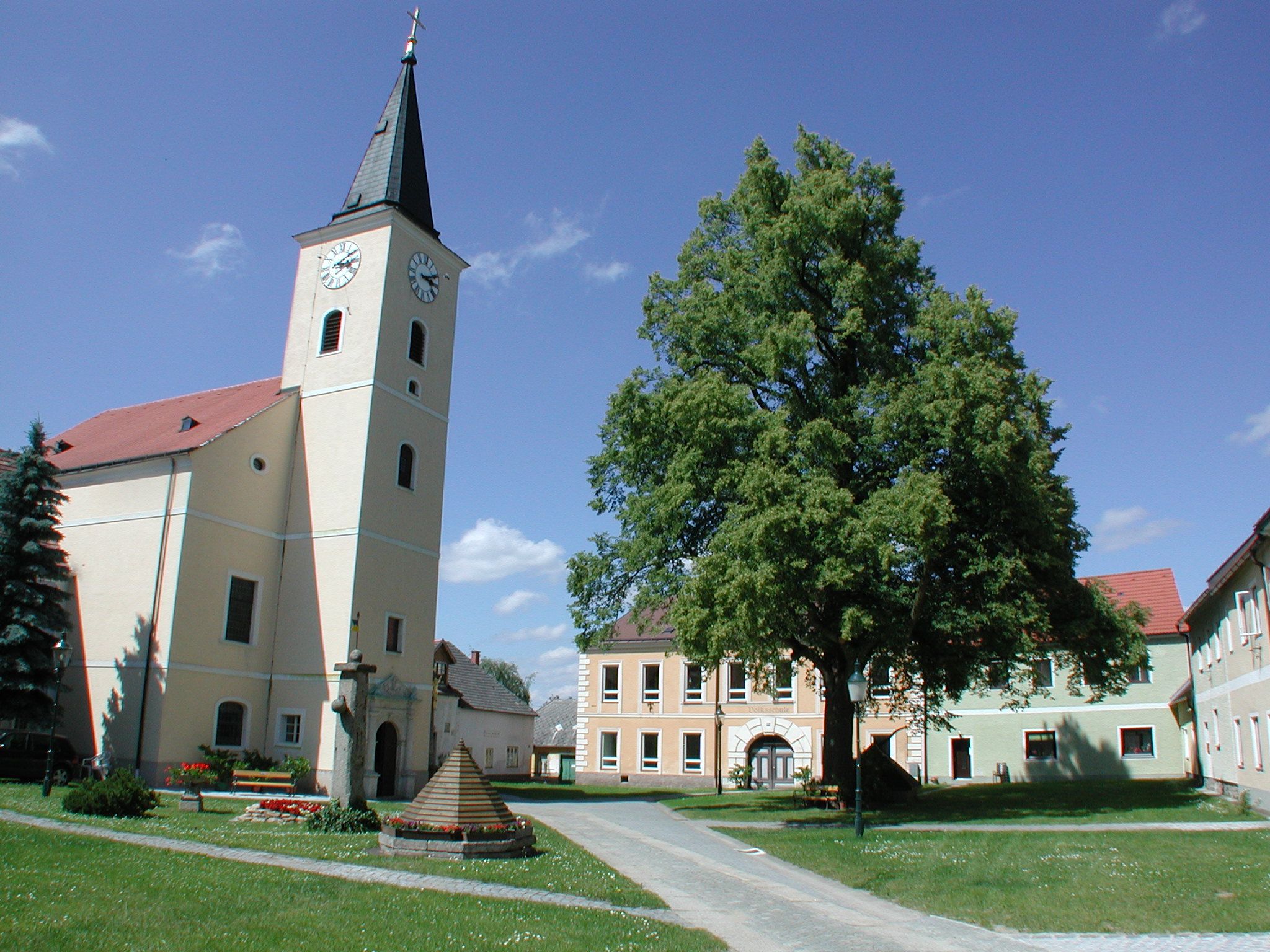 Church with tower and clock in a village square with trees and buildings.
