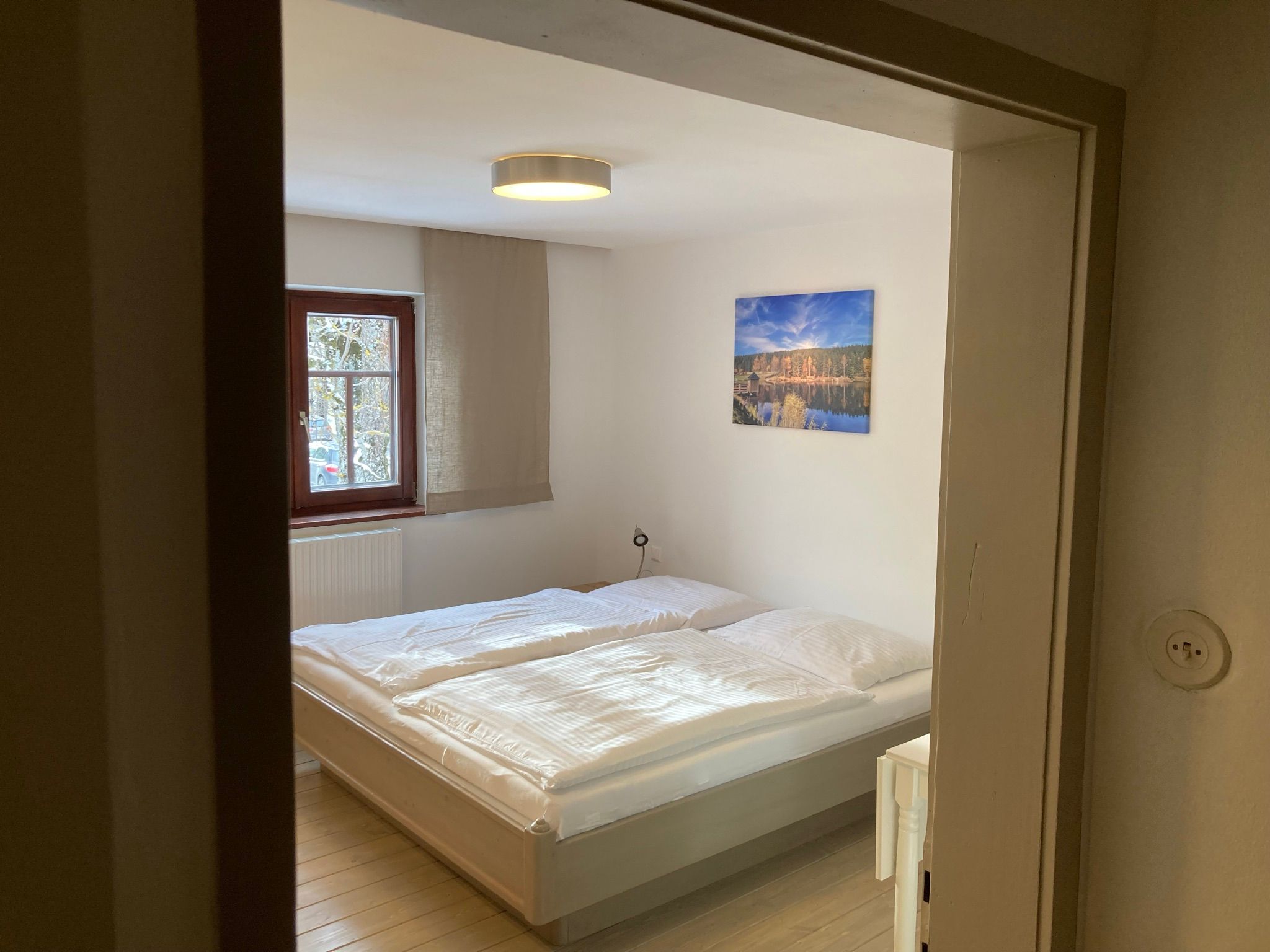 A cozy bedroom with a double bed, a window and a landscape painting on the wall.