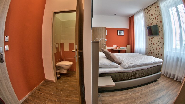 A modern guest room with double bed, desk and TV. Next to it a small bathroom with toilet.