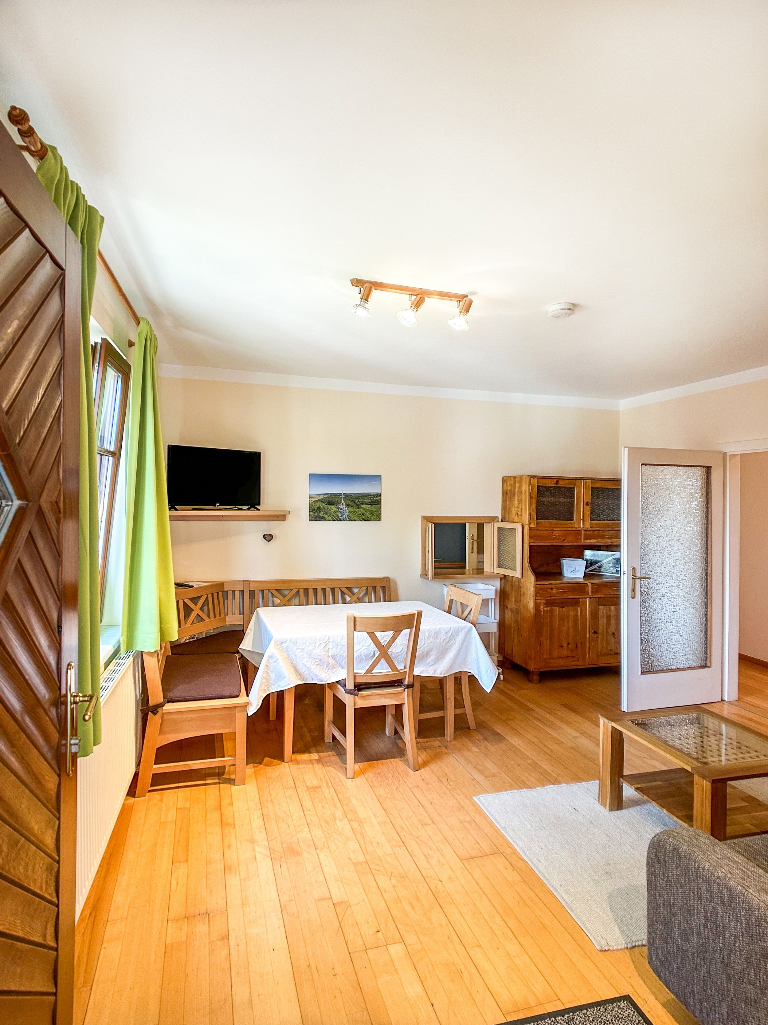 Dining room with wooden furniture, table with white tablecloth, green curtains and TV on the wall.