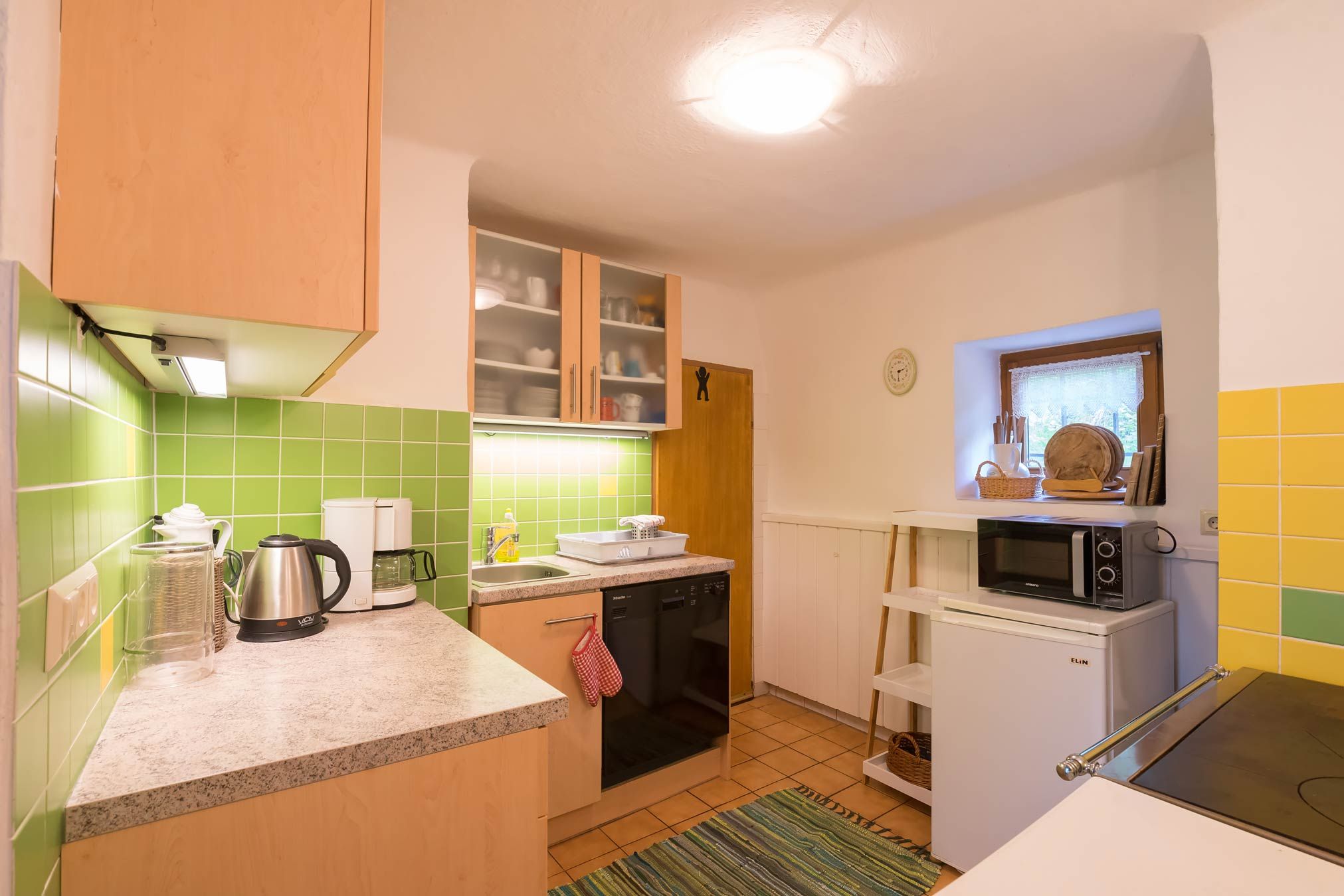 Small kitchen with green tiles, electrical appliances and wooden cupboards.