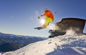 A skier jumps into the air in front of the Terzerhaus on the Gemeindealpe, surrounded by snow-covered mountains and a blue sky.