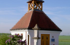 Small chapel with a red roof and cross, surrounded by flowers and a bench, under a clear sky.