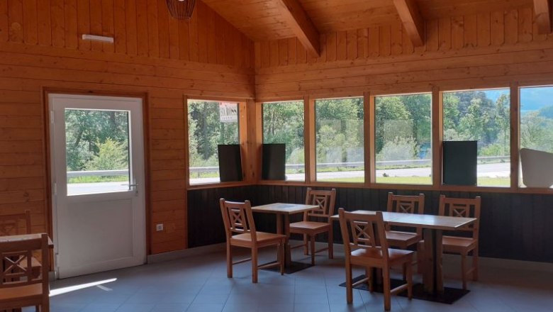 Interior view of a wooden building with tables and chairs, large windows and a door.