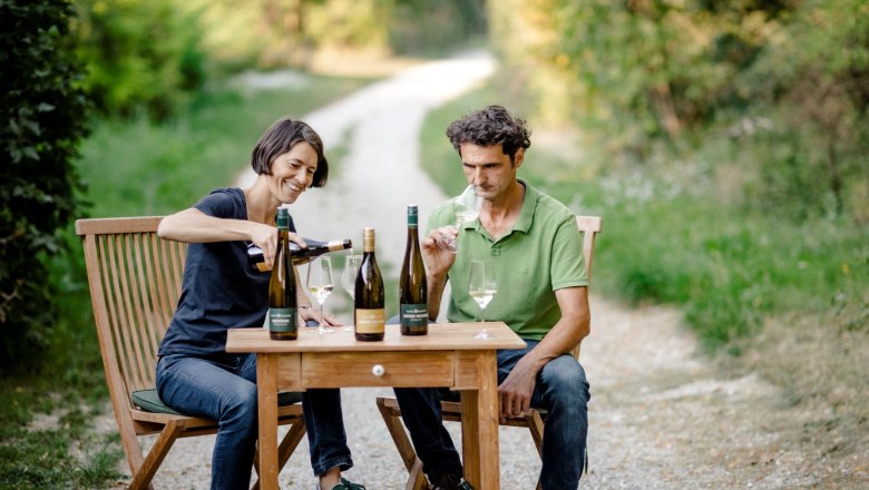 Two people sit at a wooden table outside and taste wine.