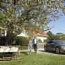 A couple walks next to a parked electric car under a blossoming tree in a rural garden.
