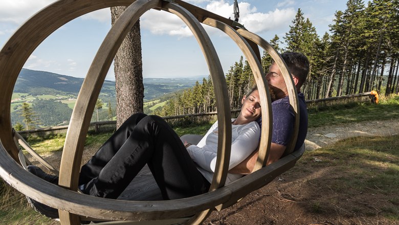 A couple relaxes in a wooden swing with a view of a hilly landscape and woods.