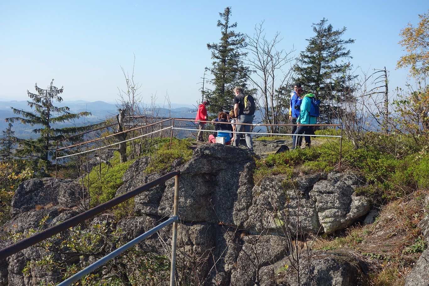 Group of hikers on a rocky viewpoint with railings, surrounded by trees and mountains in the background.