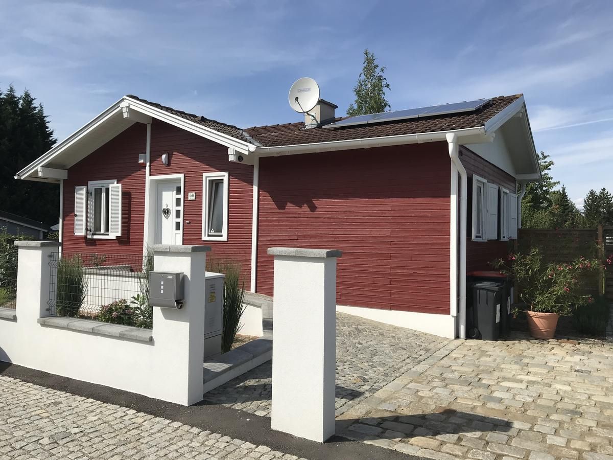 Red wooden house with white shutters and solar panels on the roof, surrounded by a paved courtyard and a low wall.
